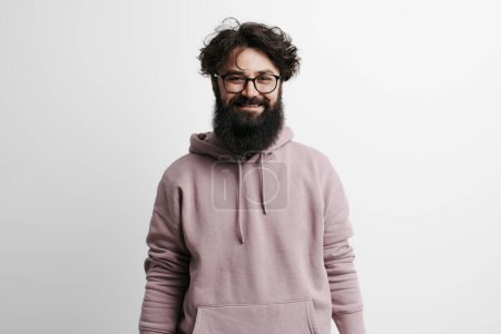 Téléchargez les photos : Homme barbu confiant avec des cheveux bouclés et des lunettes portant un sweat à capuche mauve, regardant la caméra avec un sourire amical - en image libre de droit