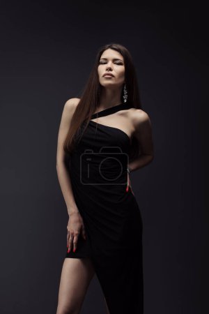 Téléchargez les photos : Un modèle époustouflant pose dans une robe noire asymétrique, son vernis à ongles rouge complétant son apparence frappante sur fond sombre - en image libre de droit