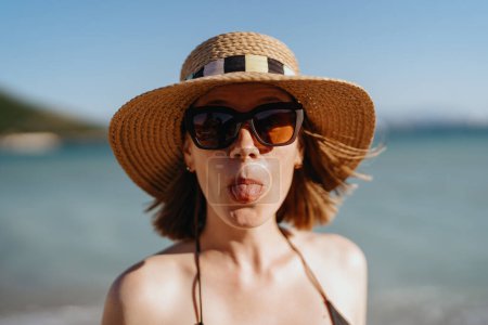 Téléchargez les photos : Une femme sort sa langue, ludique dans son chapeau et ses lunettes de soleil, avec la mer comme toile de fond - en image libre de droit