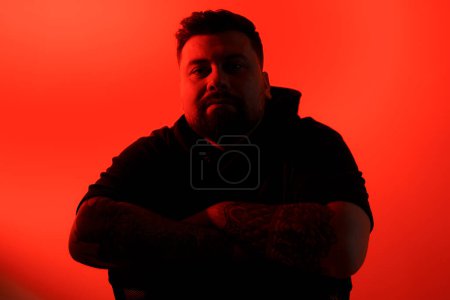Photo for Silhouetted man with arms crossed and tattoos, exuding confidence on a vibrant red background - Royalty Free Image