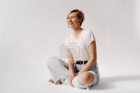 Téléchargez les photos : Femme assise jambes croisées, riant en jean denim clair et un t-shirt blanc, incarnant un esprit insouciant - en image libre de droit