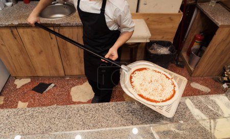 Photo for Chef in a black apron holding a pizza peel with an uncooked cheese pizza, ready for the oven - Royalty Free Image