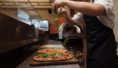 Téléchargez les photos : Un chef saupoudre délicatement de la roquette fraîche sur une pizza gourmande, ajoutant une touche de vert au plat salé - en image libre de droit