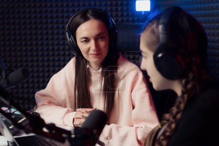 Female podcast hosts immersed in conversation, with one attentively listening, in a soundproof studio environment, indicative of a thoughtful and collaborative dialogue