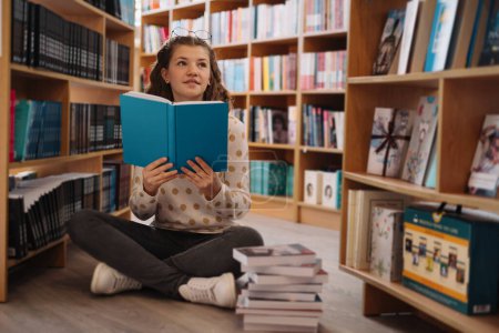 Photo for Thoughtful teenage girl with glasses on her head reads a book, seated cross-legged near a stack of books in a library - Royalty Free Image