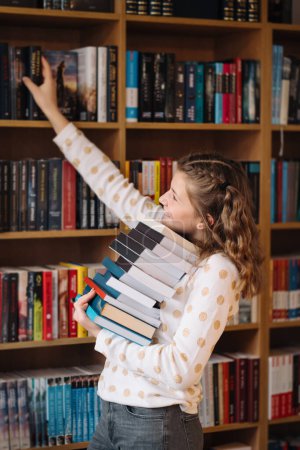Ein Teenager im gepunkteten Pullover greift nach einem Buch auf einem hohen Regal und balanciert einen großen Stapel in ihren Armen.