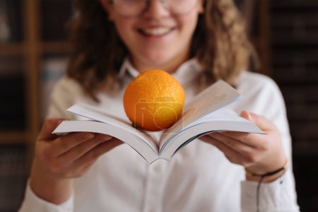 A cheerful girl balances an orange on an open book, blending learning with a healthy snack