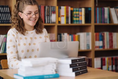Photo for Teen girl with glasses smiles while working on a laptop in the library, with piles of books nearby - Royalty Free Image