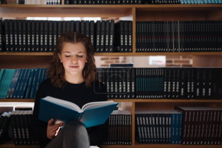 Ein fokussiertes Teenager-Mädchen liest ein blaues Buch in einer Bibliothek, umgeben von Regalen voller Literatur