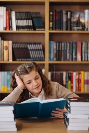 Téléchargez les photos : Une adolescente, lunettes sur la tête, est absorbée par la lecture d'un livre à la bibliothèque, entourée de piles de livres - en image libre de droit
