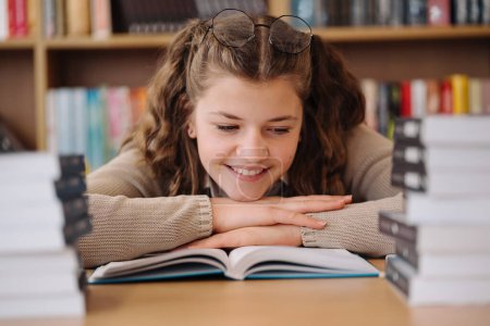 Photo for A teenage girl with glasses perched on her head is captivated by a book in the library, with a joyful expression - Royalty Free Image