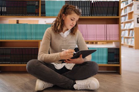 Téléchargez les photos : Une adolescente s'assoit les jambes croisées sur le sol, profondément concentrée sur sa tablette pour étudier dans un cadre de bibliothèque moderne - en image libre de droit