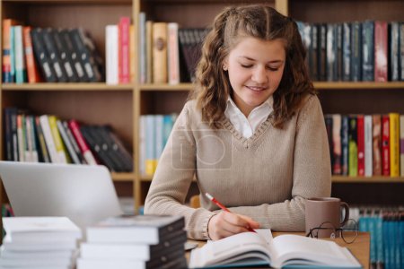Téléchargez les photos : Engrossie dans ses études, une adolescente avec un stylo et un livre travaille diligemment à une table de la bibliothèque - en image libre de droit