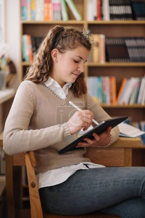 Photo for A teenage girl with glasses focused on her tablet, taking notes with a stylus, comfortably seated in the library - Royalty Free Image