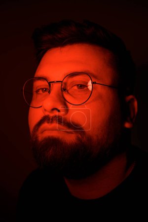 A mans contemplative expression is captured under a haunting red glow, emphasizing his glasses and beard