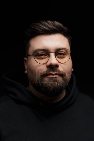 Photo for Close-up of a self-assured adult male with stylish glasses and a neat beard against a black backdrop - Royalty Free Image