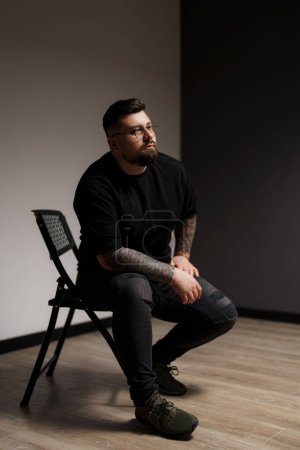 Un hombre elegante con tatuajes y gafas se sienta contemplativamente en una silla, exudando un comportamiento tranquilo y reflexivo