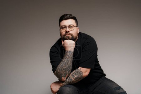 A thoughtful man with sleeve tattoos and glasses poses with his hand on his chin, looking reflective
