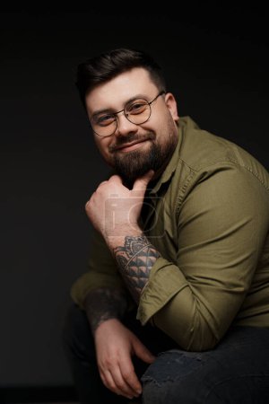 A friendly bearded man with stylish glasses smiles warmly, showcasing his sleeve tattoos in an olive shirt