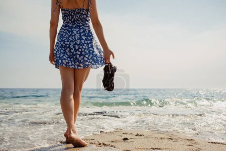 Barfuß und unbeschwert genießt eine Frau mit Sandalen in der Hand den weichen Sand und die sanften Wellen entlang der sonnigen Küste