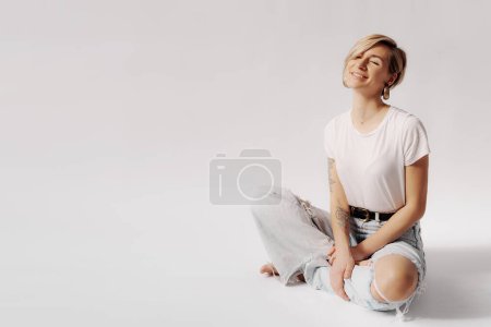 A joyful young woman sitting in a relaxed pose, smiling with eyes closed in a light denim and white t-shirt
