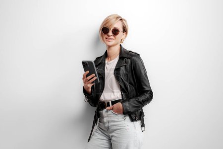 Fashionable young woman interacts with a smartphone, sporting a leather jacket and sunglasses for a tech-savvy look