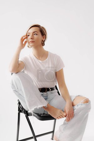 A contemplative young woman poses in a white tee and light denim, seated on a modern chair, exuding a casual elegance