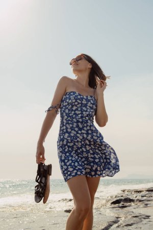 A cheerful woman in a blue dress enjoys the sun on a beach, with a gentle breeze in her hair