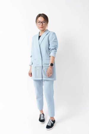 Young professional woman in a light blue suit and sneakers posing on white background.
