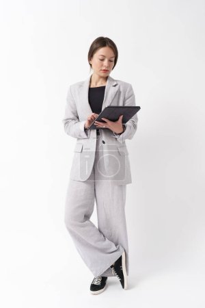 Focused businesswoman using a digital tablet, dressed in a formal suit against a white backdrop.