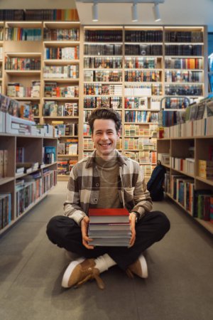 Cheerful young adult holding a stack of books in a bright, orderly library.