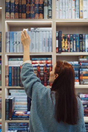 Photo for A woman stretches to pick a book from the high shelf in a bookstore. - Royalty Free Image