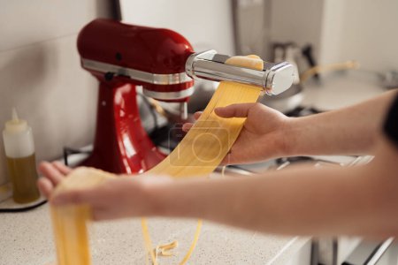 Hands preparing homemade pasta with a red pasta machine on a kitchen countertop.