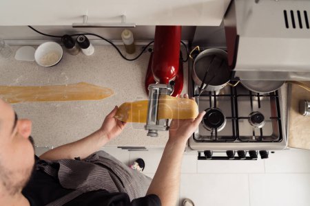Top view of person using a pasta maker to roll fresh dough in a home kitchen.