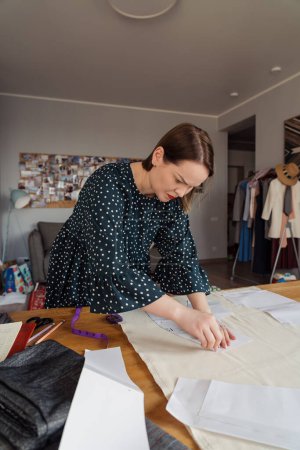 Focused fashion designer tracing patterns on fabric, surrounded by tailoring tools and garments in a creative studio setting.