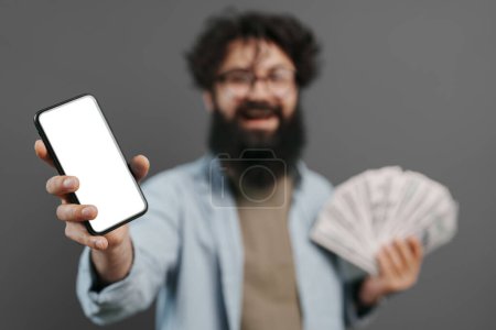 Un hombre barbudo exuberante sostiene un teléfono inteligente con una pantalla en blanco y un fan del dinero en efectivo, expresando el éxito y las oportunidades de finanzas digitales contra un fondo gris neutro.