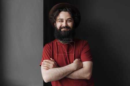 Retrato de un hombre barbudo alegre con una sonrisa amistosa, con una camiseta roja y un sombrero elegante, posando con confianza con los brazos cruzados sobre un fondo gris.