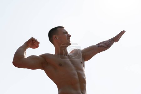 A shirtless muscular man with tattoos flexes his biceps and smiles confidently against a clear sky backdrop, radiating strength and positivity.