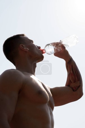 A shirtless athletic man with a tattoo on his arm hydrates by drinking water from a clear bottle against a bright sky backdrop. The image conveys concepts of fitness, health, and hydration.