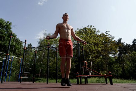 A young man with a toned physique trains at an outdoor fitness park, surrounded by green trees under a clear sky. He stands confidently, showcasing strength and dedication on the workout bars.