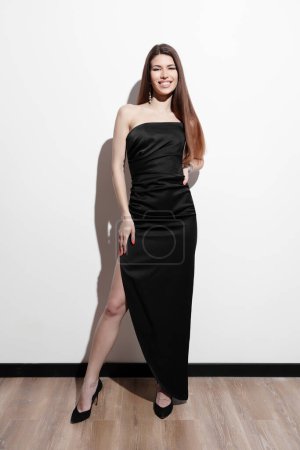 A poised and elegant woman in a chic black evening dress stands confidently against a white wall, exuding class and a timeless style. Perfect for themes of elegance, fashion, and confidence.