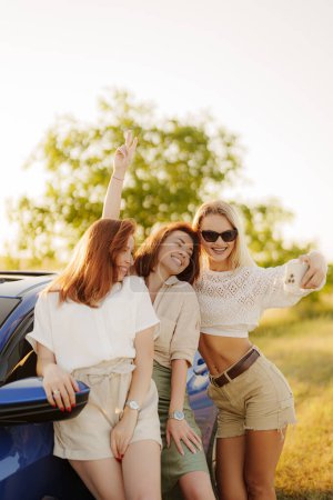 Group of cheerful young friends posing for a selfie beside a car on a sunny summer day, capturing memories of their road trip.
