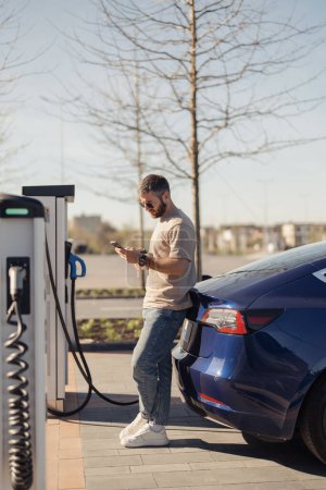 Casual man using smartphone during the charging of his electric vehicle at a public charging station on a sunny day.