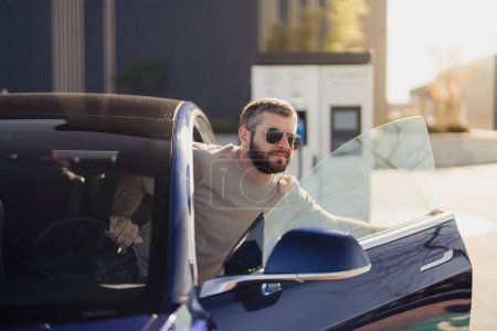 Stilvoller bärtiger Mann mit Sonnenbrille steigt aus einem modernen blauen Elektroauto und präsentiert umweltfreundlichen Transport.