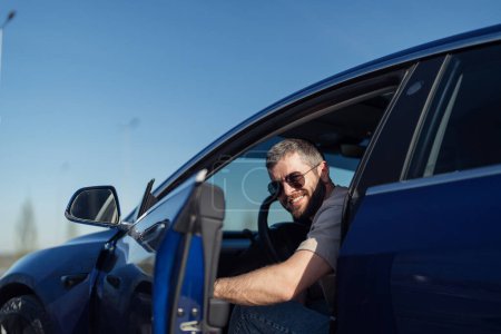 Porträt eines fröhlichen, bärtigen Mannes mit Sonnenbrille, der sich aus seinem neuen Elektroauto lehnt und einen sonnigen Tag mit strahlend blauem Himmel genießt.