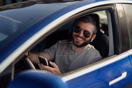 Foto de Hombre sonriente con gafas de sol usando su teléfono inteligente mientras está sentado en el asiento del conductor de un coche eléctrico moderno. - Imagen libre de derechos