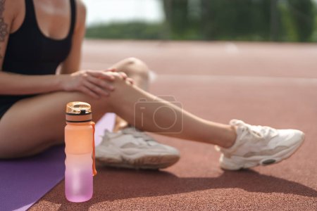 Primer plano de una atleta sentada en una esterilla de yoga en una pista de atletismo, descansando con una botella de agua enfocada.