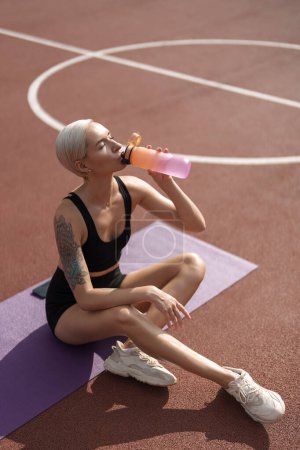 Fitness enthusiast taking a break to drink water on a sunny day at the athletic track with yoga mat.