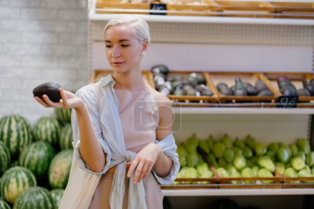 Una joven compradora examina un aguacate con una expresión reflexiva en la sección de productos de una tienda de comestibles, rodeada de frutas frescas.