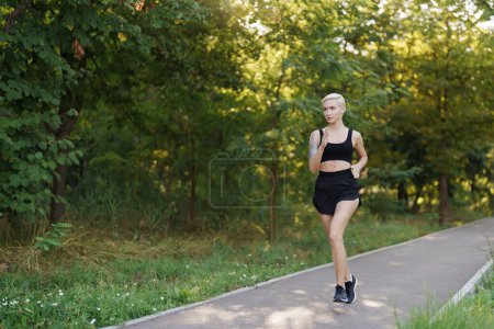 A fit young woman with a pixie haircut is shown jogging on a scenic park path surrounded by lush green trees, conveying health and vitality.